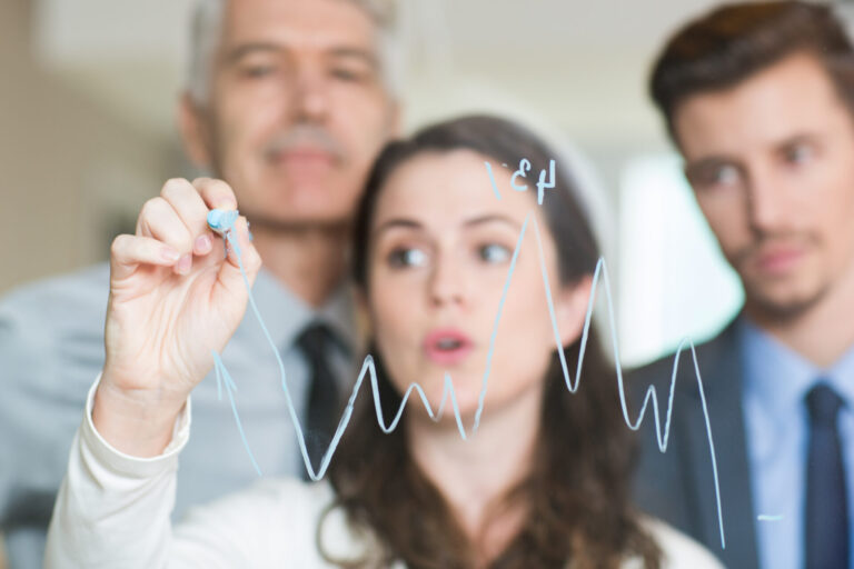 Uma mulher de negócios ilustra um gráfico de linhas ascendentes com um marcador em um vidro transparente, demonstrando o crescimento possível com um software para lotéricas, enquanto dois colegas homens observam atentamente ao fundo.