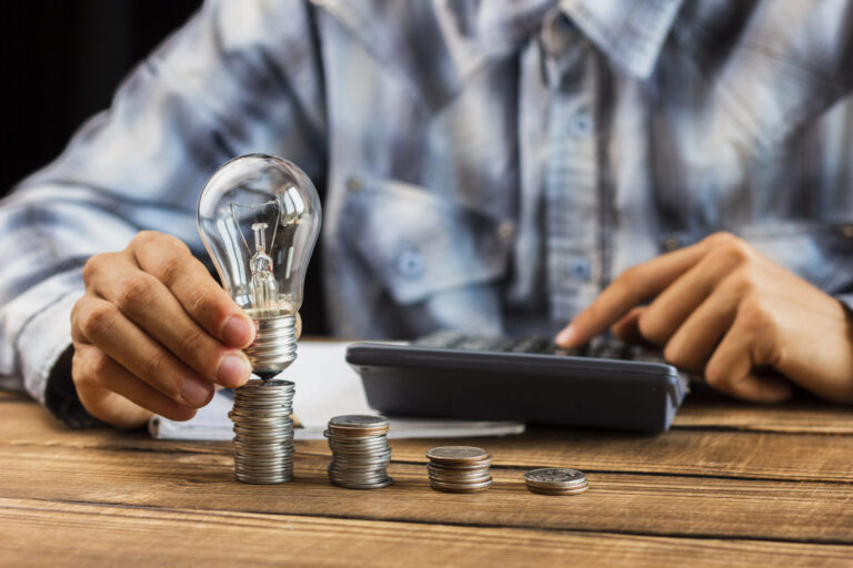 Uma pessoa de camisa xadrez empilha moedas em uma mesa de madeira, segurando uma lâmpada em cima de uma pilha enquanto usa uma calculadora com a outra mão - simbolizando economia inteligente e eficiência, como um sistema para lotéricas.