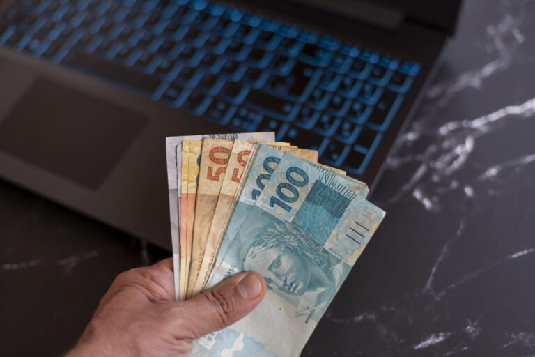 Uma mão segura várias cédulas de real de diferentes valores em frente a um laptop preto com teclas azuis retroiluminadas em uma superfície de mármore escuro, sugerindo transações possivelmente gerenciadas por um software para lotéricas.