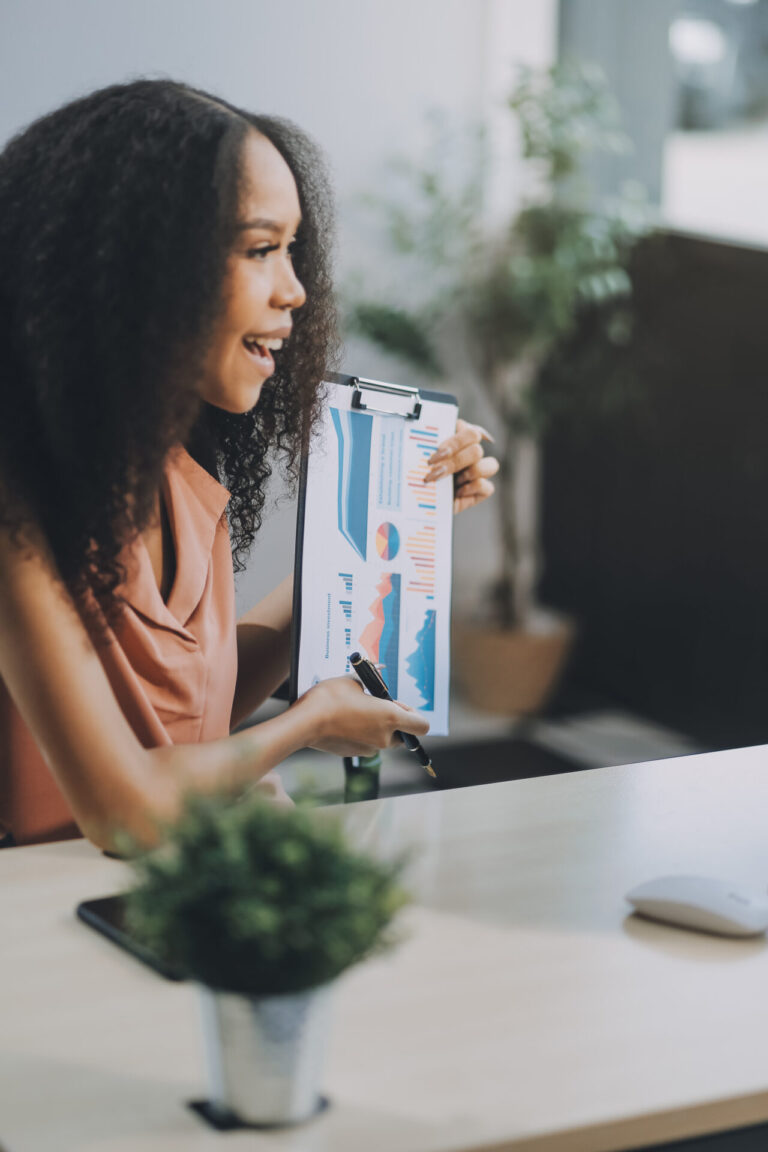 Uma mulher sentada em uma mesa, sorrindo e segurando uma prancheta com gráficos coloridos, apontando para dados de um software para lotéricas durante uma apresentação. Um pequeno vaso de planta dá um toque de verde à mesa.