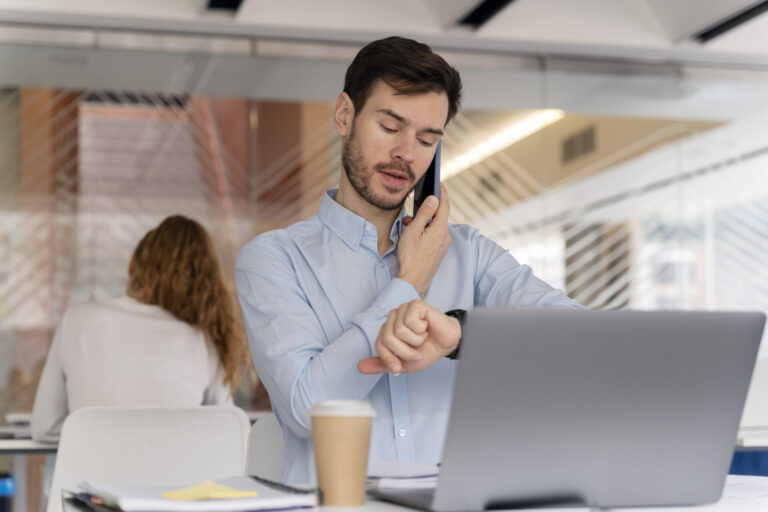 Um homem de camisa azul está sentado em uma mesa, falando ao telefone e verificando o relógio. Um laptop com software para lotéricas, uma xícara de café e um bloco de notas estão sobre a mesa. Outra pessoa trabalha em segundo plano.