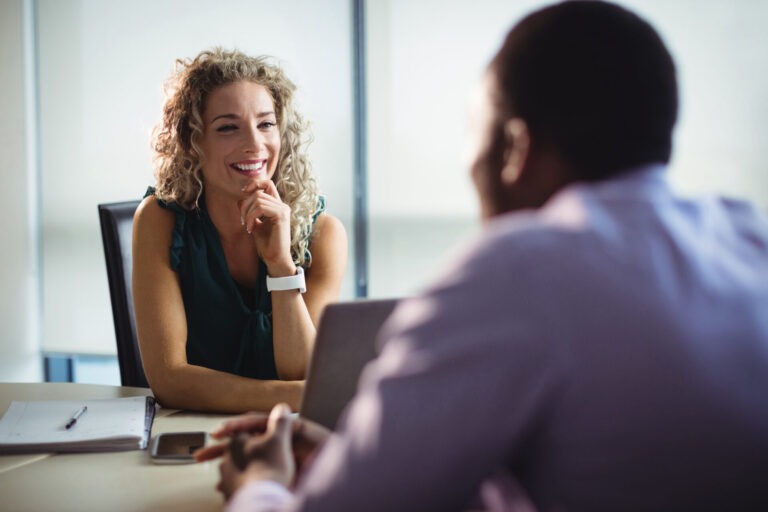 Uma mulher de cabelos cacheados sorri e ouve durante uma conversa com um homem em uma mesa de escritório, possivelmente discutindo os benefícios de um software para lotéricas. Um notebook, um smartphone e papéis estão sobre a mesa.