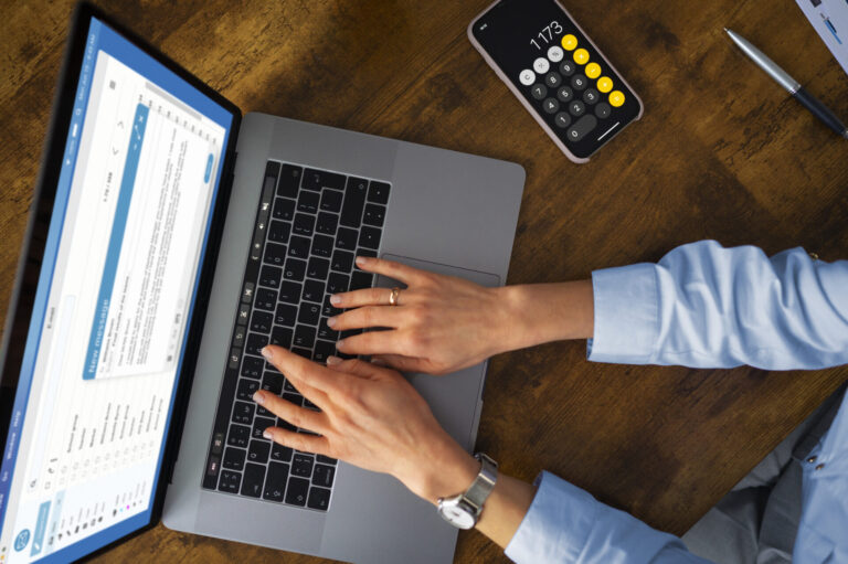 Uma pessoa digita em um laptop que exibe uma caixa de entrada de e-mail, possivelmente usando um software para lotéricas. Um smartphone com um aplicativo de calculadora mostrando "1173" e uma caneta estão sobre a mesa de madeira ao lado de uma pessoa com camisa azul clara e relógio.
