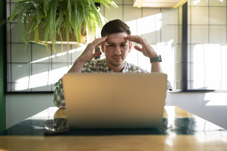 Um homem senta-se em uma mesa em frente a um laptop, tocando as têmporas e parecendo estressado enquanto tenta gerenciar tarefas sem um sistema para lotéricas. Um smartphone está sobre a mesa e uma planta frondosa está pendurada ao fundo.