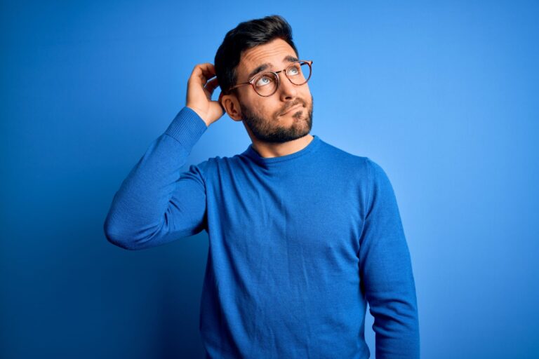 Um homem com cabelos escuros curtos, barba e óculos está de pé contra um fundo azul, vestindo um suéter azul. Ele coça a cabeça com uma expressão confusa, como se estivesse ponderando sobre o melhor software para lotéricas para suas necessidades.