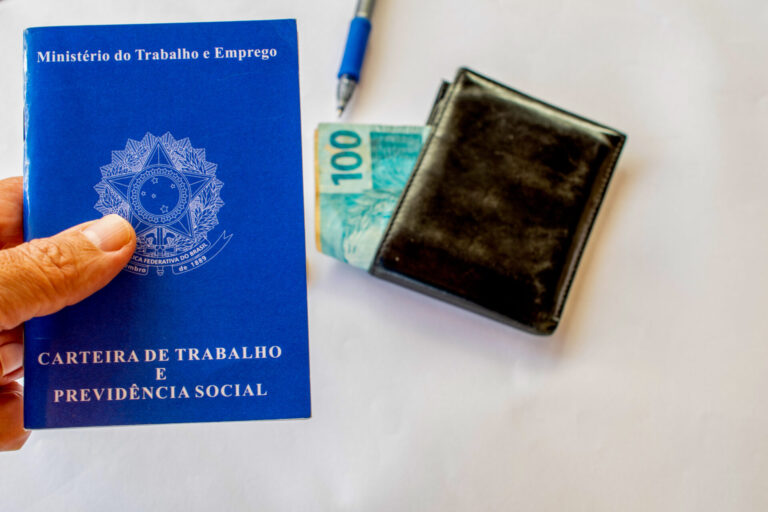 Uma mão segura uma carteira de trabalho e previdência social brasileira ao lado de uma carteira preta com uma nota de 100 reais, ilustrando a importância de um software para lotéricas. Uma caneta está parcialmente visível em uma superfície branca.