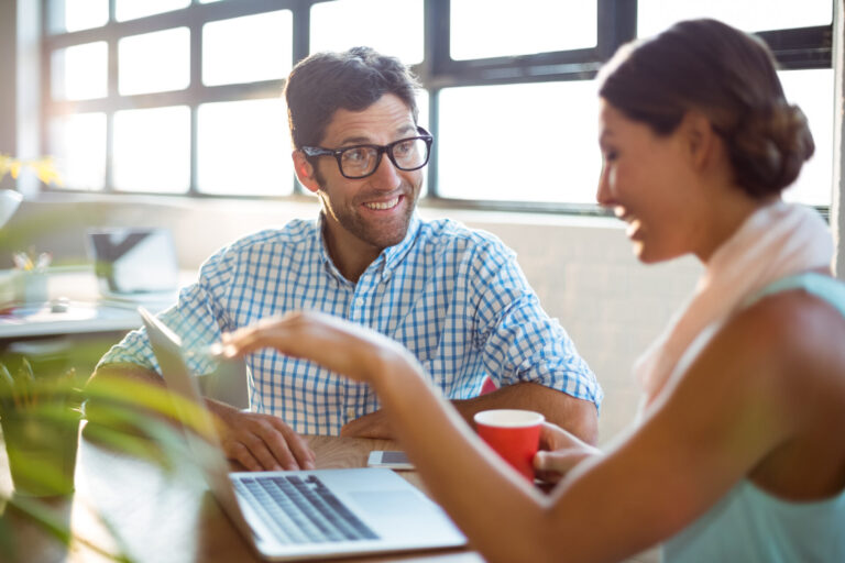 Duas pessoas sentadas em uma mesa com um laptop, sorrindo e conversando sobre um software para lotéricas. Uma delas segura uma caneca vermelha enquanto gesticula, e a outra, usando óculos, ouve atentamente enquanto a luz do sol atravessa as grandes janelas atrás delas.