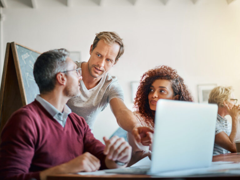 Três pessoas sentadas em uma mesa com um laptop, discutindo um sistema para lotéricas enquanto uma delas se levanta e aponta para a tela. Os outros ouvem atentamente, com um quadro-negro e outra pessoa visíveis ao fundo.