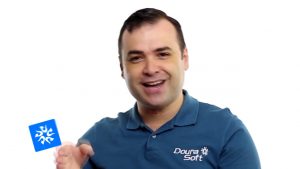 Um homem de cabelos escuros curtos, sorrindo e gesticulando com uma das mãos. Ele está vestindo uma camisa polo azul com o logotipo da Doura Soft, pronto para ajudar você a vender meus clientes. O plano de fundo é branco simples, com um pequeno ícone azul no canto inferior esquerdo.