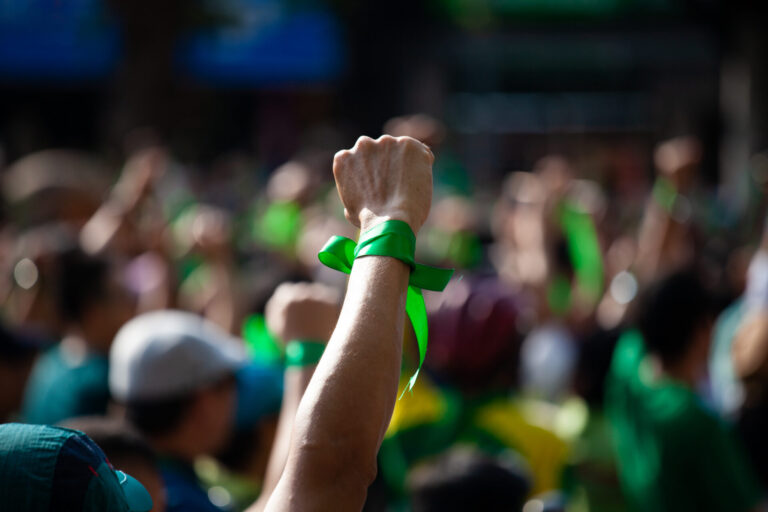 Um punho levantado com uma fita verde amarrada ao redor do pulso se destaca em meio a uma multidão de pessoas, muitas delas vestindo verde, em um ambiente externo, simbolizando unidade ou protesto - muito parecido com as cenas discutidas no informe febralot.
