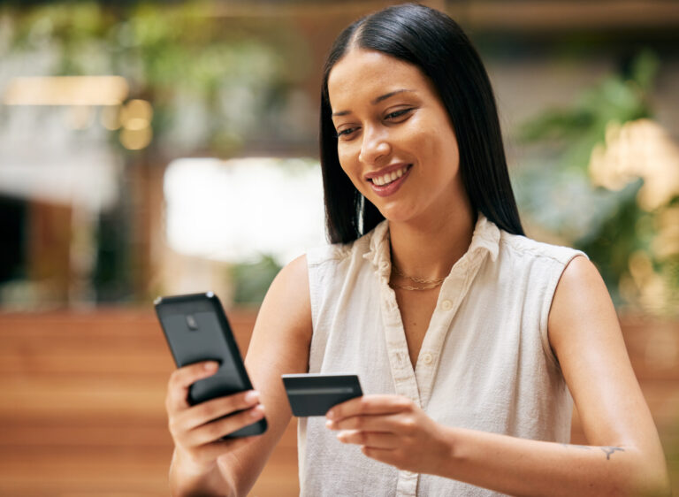Uma jovem com longos cabelos escuros sorri enquanto segura um smartphone em uma das mãos e um cartão de crédito na outra, parecendo fazer uma compra on-line enquanto novas regras do cartão de crédito passam a valer em um ambiente interno iluminado.