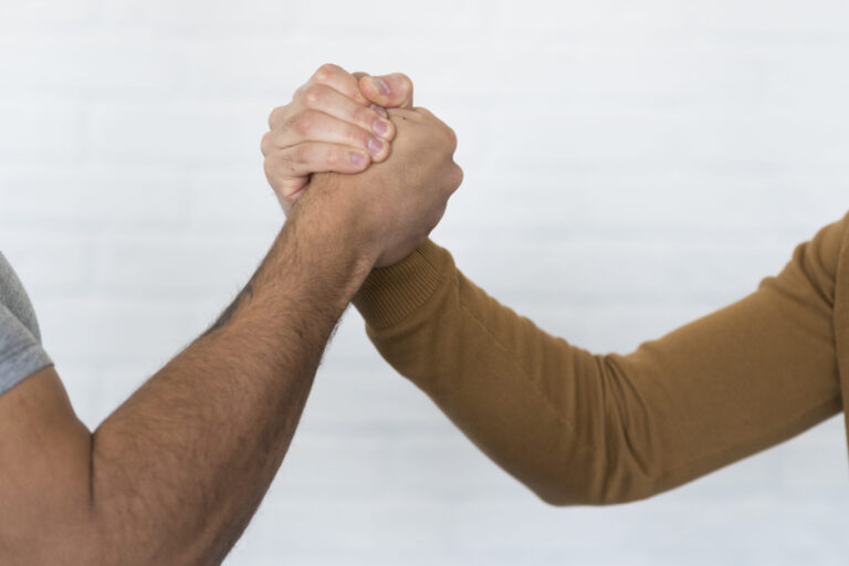 Duas pessoas estão de mãos dadas em uma pose de aperto de mão ou de braço de ferro contra um fundo claro e desfocado, simbolizando parceria e confiança - valores essenciais ao escolher um software para lotéricas. Um usa uma manga marrom e o outro uma manga curta cinza.