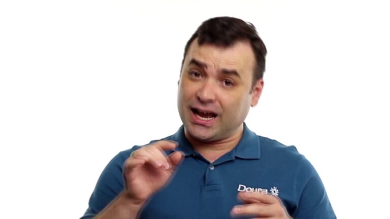Um homem de cabelos escuros vestindo uma camisa polo azul está falando e gesticulando com as mãos, demonstrando um comportamento confiante e negocial. O plano de fundo é branco puro.