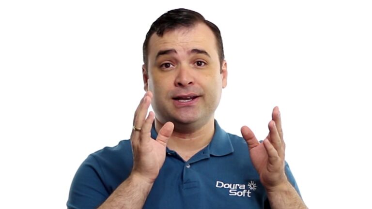 Um homem vestindo uma camisa polo azul da Doura Soft gesticula com as duas mãos enquanto fala sobre o Gerador de relatórios. Ele tem cabelos escuros curtos e está de pé contra um fundo branco liso.