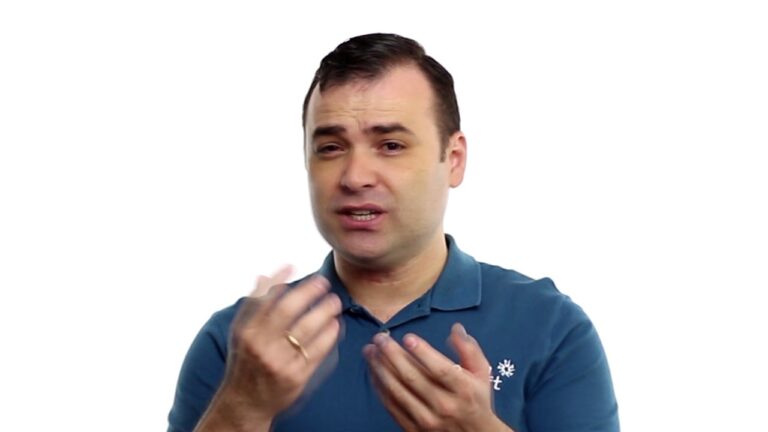 Um homem vestindo uma camisa polo azul gesticula com as mãos enquanto fala sobre a Gestão Verdadeira. Ele tem cabelos escuros curtos e está de pé contra um fundo branco liso.