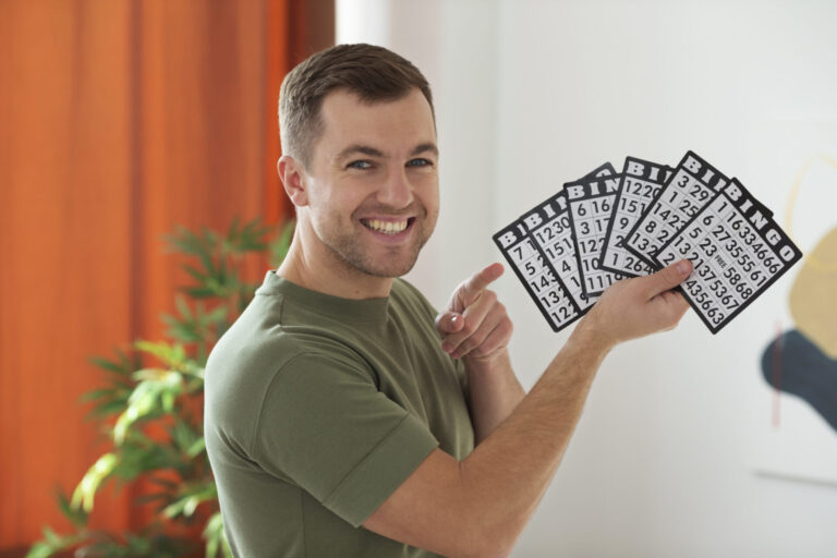 Um homem sorridente, de camisa verde, segura várias cartelas de bingo em uma mão e aponta para elas com a outra, em um ambiente interno próximo a uma planta e cortinas laranja, enquanto as notícias dizem que a comissão rejeita a legalização de bingos para arrecadação.