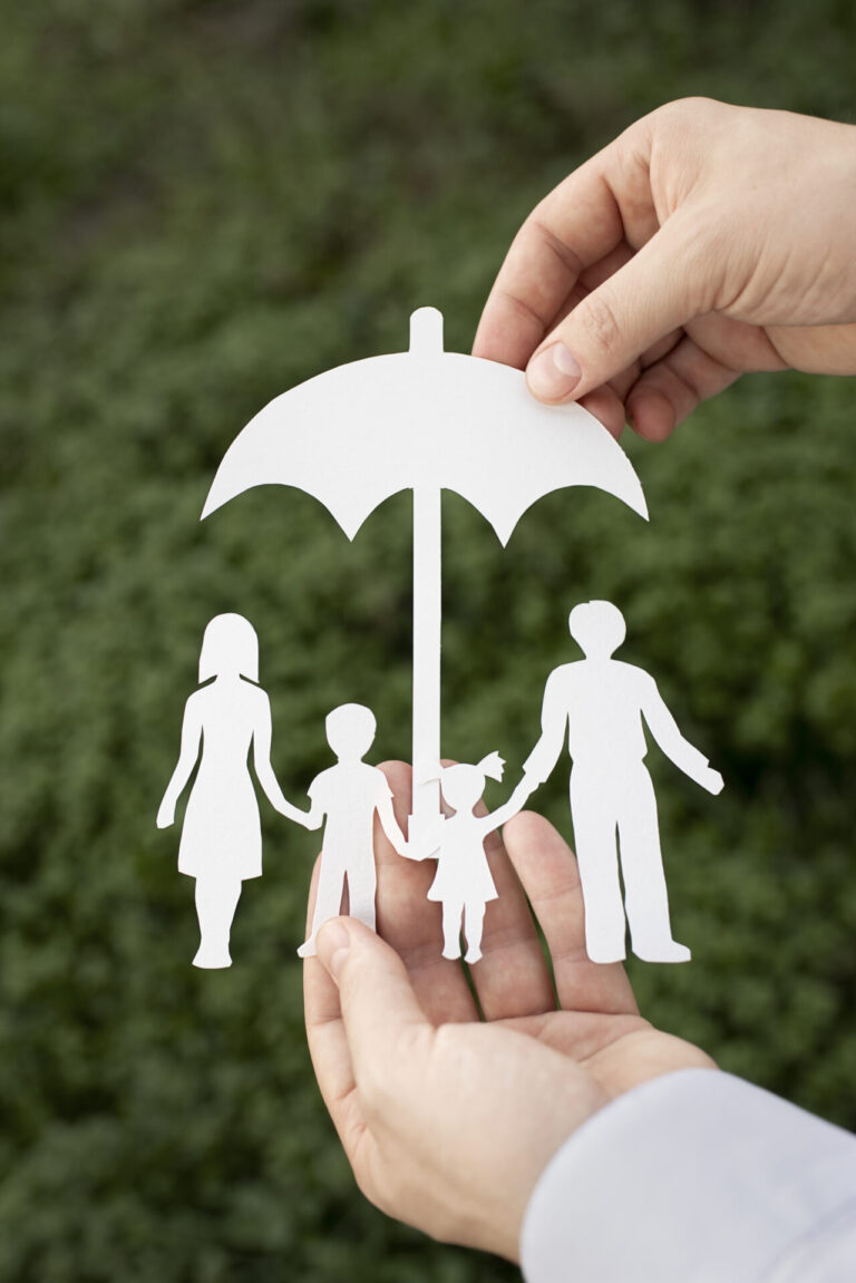 Duas mãos seguram um recorte de papel de uma família de quatro pessoas sob um guarda-chuva, simbolizando proteção - assim como o seguro vida da gente - com um fundo natural verde e desfocado.
