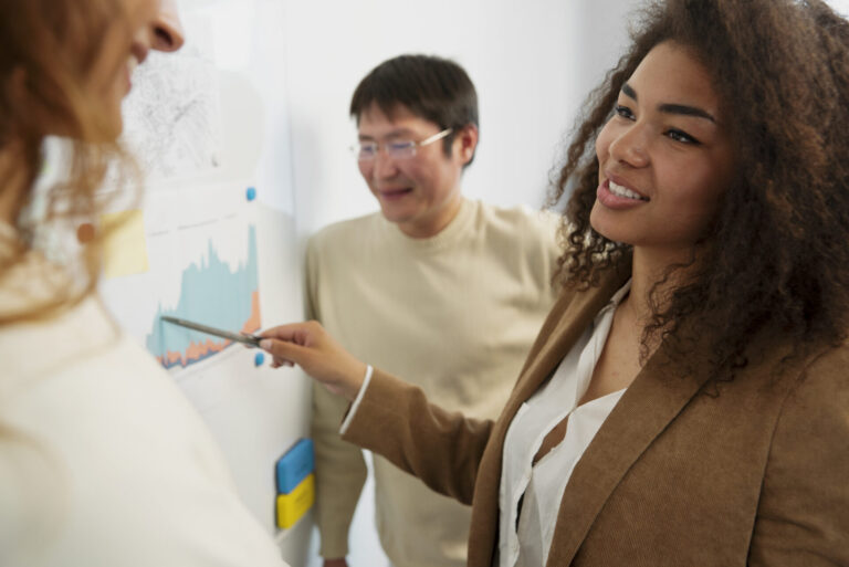 Uma mulher de blazer marrom aponta para um gráfico em um quadro branco, explicando os benefícios de um sistema para lotéricas enquanto sorri e conversa com dois colegas durante uma reunião casual no escritório.