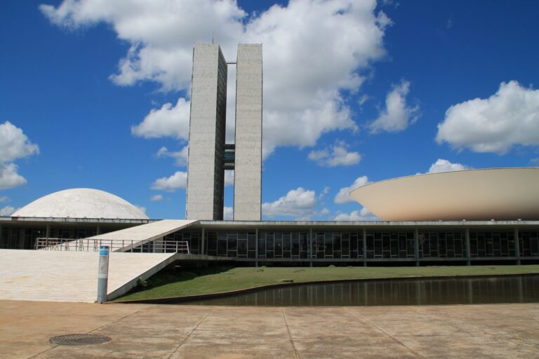 Um edifício governamental moderno com duas torres retangulares altas, uma estrutura em cúpula branca à esquerda e uma estrutura em forma de tigela à direita, frequentemente apresentado nas atualizações de notícias do comunicado à rede - aprovação pl 7306/2017, em um céu azul com nuvens dispersas.