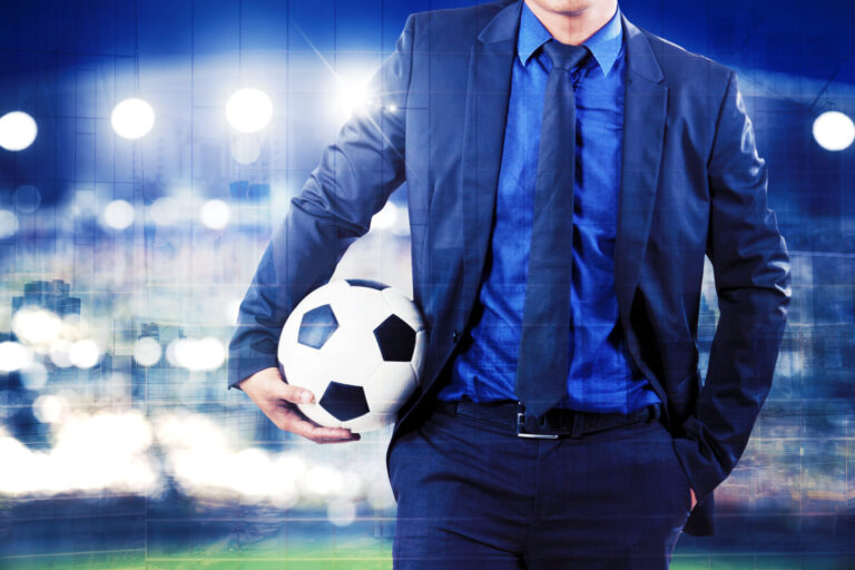 Um homem de terno escuro e camisa azul segura uma bola de futebol debaixo do braço, em um estádio bem iluminado - talvez onde os projetos que legalizam jogos de azar estejam gerando debates acalorados entre torcedores e espectadores.