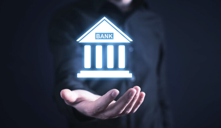 Uma pessoa de camisa escura estende a mão com um ícone virtual brilhante de um prédio de banco pairando sobre sua palma, simbolizando o banco digital e como os bancos aprovam a contribuição sindical no mundo em evolução da tecnologia financeira.