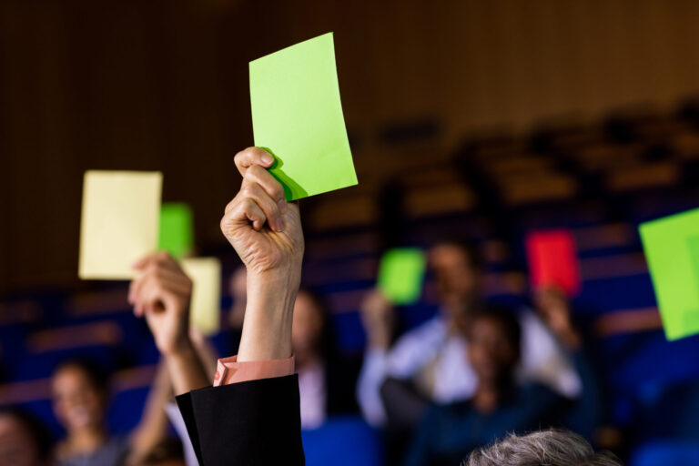 Os membros da plateia seguram cartões coloridos, principalmente verdes e amarelos, em uma sala de aula com assentos azuis, enquanto o jogo avança no país à espera durante uma atividade envolvente de votação ou tomada de decisão em grupo.