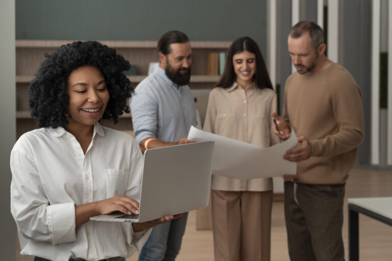 Uma mulher de camisa branca sorri enquanto usa um laptop com software para lotéricas em primeiro plano; atrás dela, três colegas discutem grandes folhas de papel em um ambiente de escritório moderno.