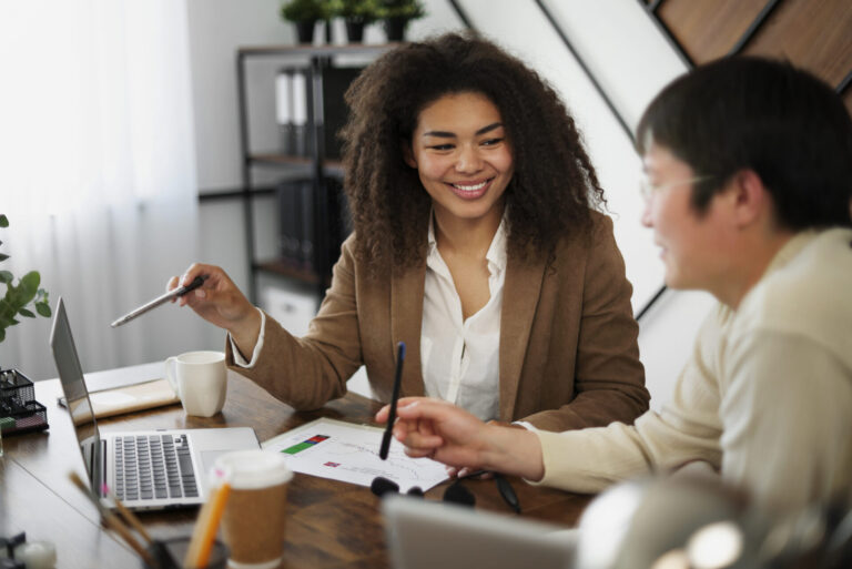 Dois colegas sentados em uma mesa com laptops e papéis, sorrindo enquanto discutem um novo software para lotéricas. Um deles faz gestos com uma caneta enquanto o outro ouve. Copos de café e material de escritório estão visíveis na mesa de madeira.