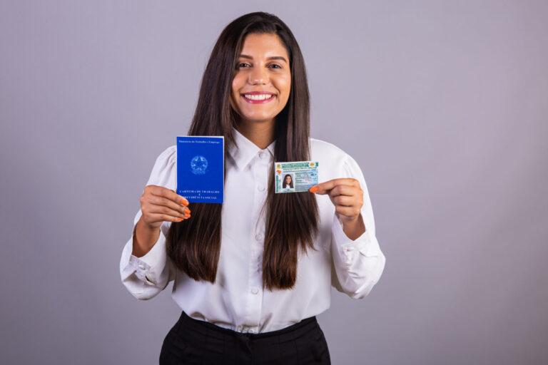 Uma mulher com longos cabelos escuros, vestindo uma blusa branca, sorri enquanto segura um passaporte azul aberto em uma das mãos e uma carteira de identidade na outra, em um fundo cinza - o governo diz que a nova lei trabalhista é importante para cidadãos como ela.