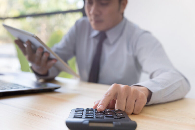 Uma pessoa em trajes de negócios está sentada em uma mesa, usando uma calculadora com uma das mãos e segurando um tablet com a outra - talvez para fazer contabilidade sindical. Um laptop está aberto nas proximidades, com a luz do dia entrando por uma janela.