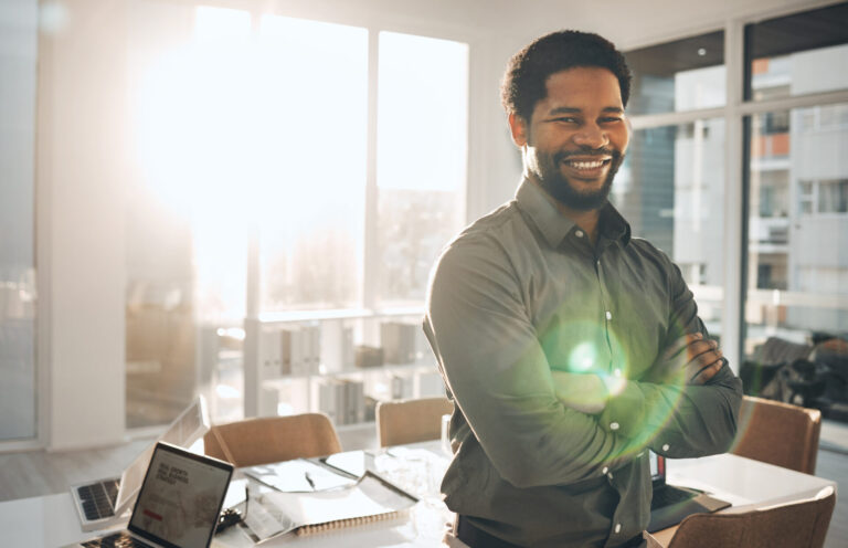 Um homem sorri com os braços cruzados em um escritório moderno e bem iluminado, com a luz do sol entrando pelas grandes janelas - um líder confiante pronto para manter uma gestão sindical eficiente. Em primeiro plano, vê-se uma mesa com um laptop, documentos e óculos.