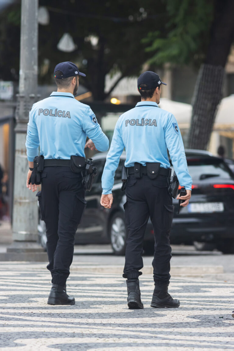 Dois policiais com camisas azuis claras e calças pretas caminham lado a lado em uma faixa de pedestres, de costas para a câmera. A palavra Polícia está visível em suas camisas, destacando o papel da segurança pública que receberá recursos de loterias em breve.