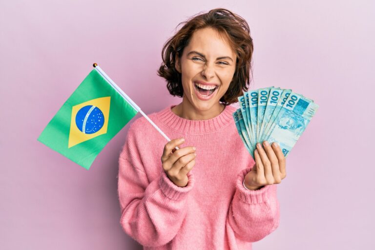 Uma pessoa sorridente, vestindo um suéter rosa, segura uma bandeira do Brasil e várias notas de 100 reais, contra um fundo rosa claro - uma cena que a quina de são joão traz à tona com sonhos de vitória.