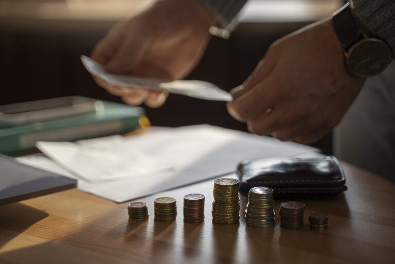 Uma pessoa em pé em uma mesa segurando papéis, com pilhas de moedas, uma carteira e envelopes em primeiro plano, ilustrando como funcionam as finanças e um orçamento ou planejamento financeiro eficaz andam de mãos dadas.