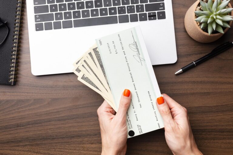 Uma pessoa com unhas pintadas de laranja segura um contracheque e várias notas de dólar americano sobre uma mesa de madeira com um laptop, um notebook, uma caneta e um pequeno vaso de suculentas, refletindo regras de movimentação de cheques conta 043.