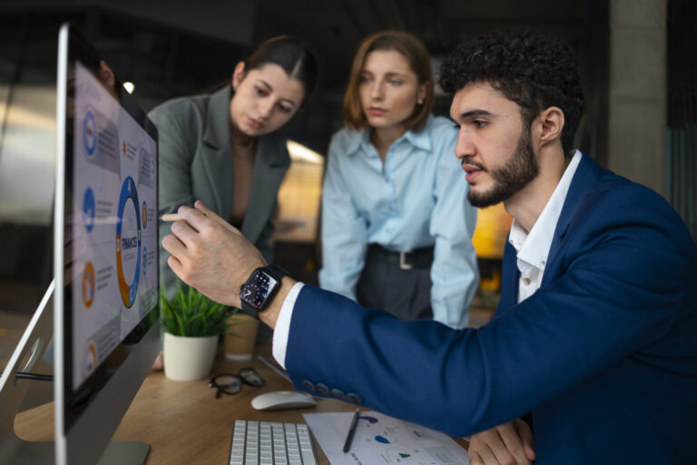Três pessoas em trajes de negócios olham para um monitor de computador que exibe gráficos com detalhes precisos. Um homem está apontando para a tela enquanto os outros dois observam atentamente. Há papéis e uma planta sobre a mesa.