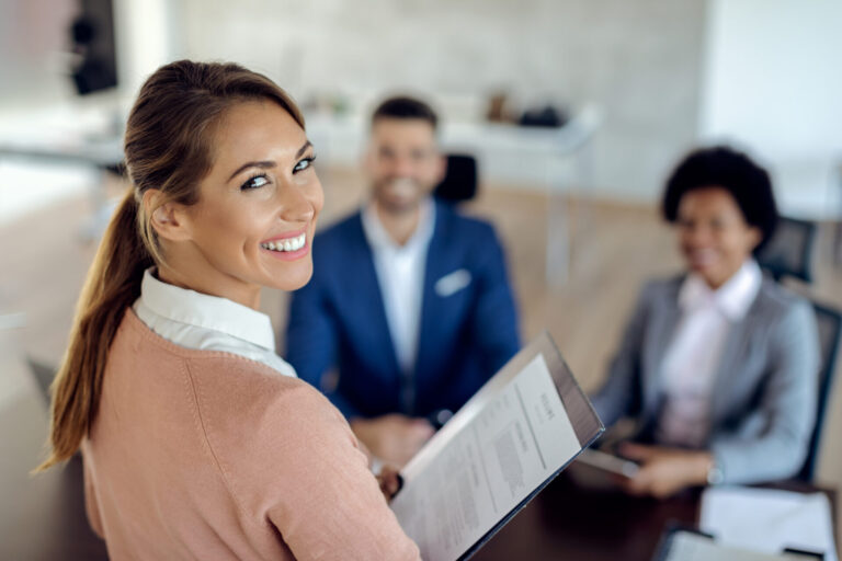 Uma mulher sorridente segurando um currículo olha para a câmera enquanto duas pessoas se sentam em frente a ela em uma mesa, sugerindo um cenário de entrevista de emprego em um escritório durante o seminário CIBELAE e Caixa.