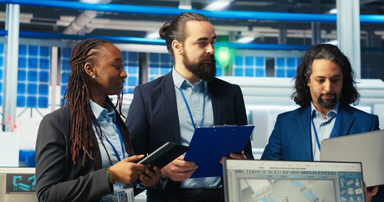 Três pessoas em trajes de negócios discutem o trabalho em um escritório moderno com monitores de computador, iluminação azul e diagramas técnicos; elas analisam detalhes enquanto a Caixa abre processo seletivo para vice-presidentes. Um deles segura um tablet, outro uma prancheta e o terceiro usa um laptop.