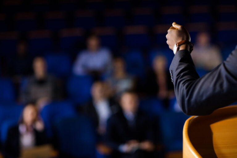 Uma pessoa de terno está em um pódio, gesticulando com a mão enquanto discute a reforma, dirigindo-se a um público desfocado sentado em um auditório com cadeiras azuis. O foco está na mão e no braço do orador.