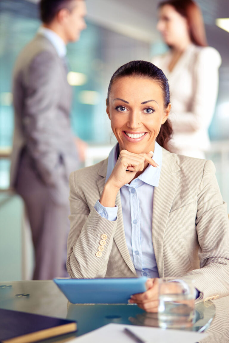 Uma mulher sorridente em um terno bege está sentada em uma mesa com um tablet e um copo de água, pronta para oferecer ajuda no cadastro de clientes. Dois colegas desfocados em trajes de negócios estão de pé e conversando ao fundo.