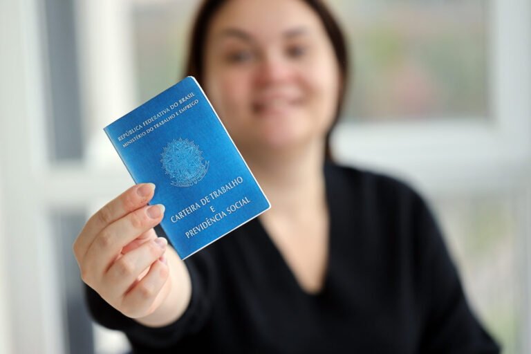 Uma mulher de camisa preta segura uma Carteira de Trabalho e Previdência Social azul, simbolizando a importância das relações trabalhistas e sindicais nas empresas. Seu rosto está fora de foco, enquanto o cartão se destaca claramente em primeiro plano.