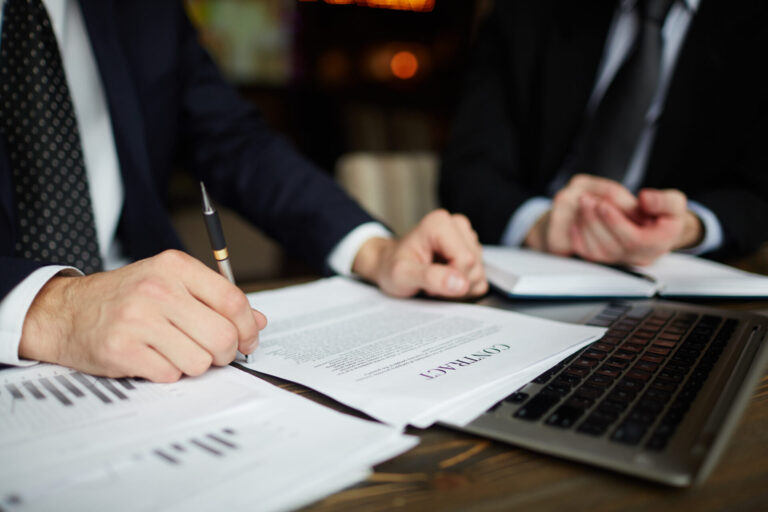 Duas pessoas em ternos de negócios sentam-se em uma mesa, analisando documentos intitulados CONTRATO sobre como a Caixa irá vender participações em loterias, com gráficos e um laptop à sua frente - uma segurando uma caneta, a outra gesticulando enquanto discutem os termos.