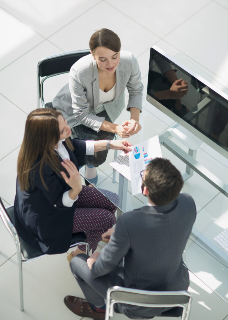 Três pessoas em trajes de negócios sentam-se ao redor de uma mesa com um monitor de computador, discutindo tabelas e gráficos impressos. A imagem, tirada de cima, destaca a relação pessoal e profissional dentro de um ambiente colaborativo.