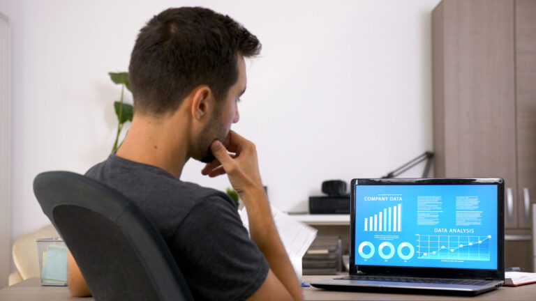 Um homem sentado em uma mesa, olhando atentamente para um laptop que exibe gráficos e tabelas com o título Dados da empresa, talvez procurando os 5 sinais de que sua lotérica não está alcançando o desempenho esperado, em um ambiente de escritório moderno e bem iluminado.