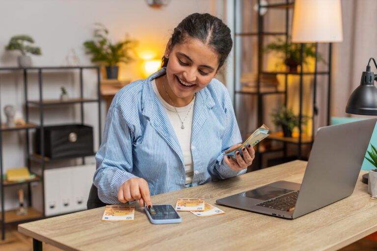 Uma mulher sorridente está sentada em uma mesa com um laptop, fazendo o fechamento de caixa da sua empresa, segurando notas em euros e usando uma calculadora, rodeada por mais cédulas em um ambiente acolhedor e bem iluminado.