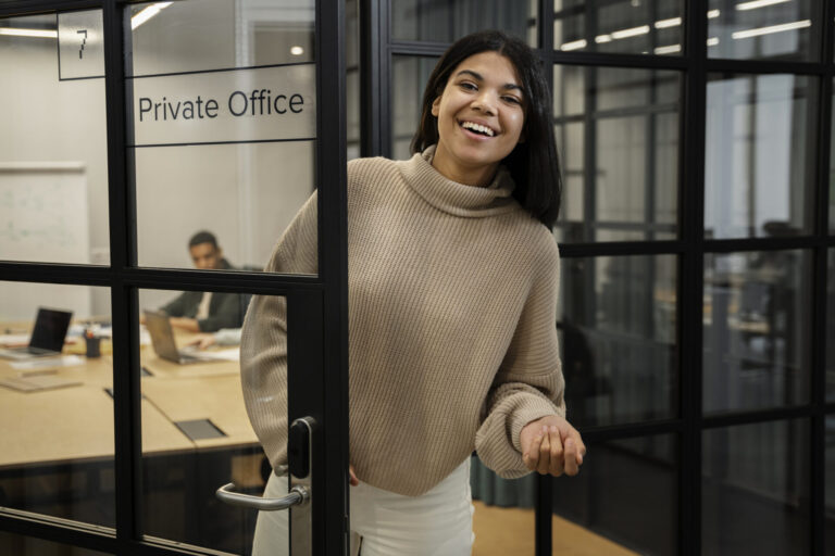 Uma mulher sorridente, vestindo um suéter bege, está na porta de um escritório particular com paredes de vidro, exibindo dicas para melhorar o ambiente físico. Lá dentro, alguém trabalha em um laptop em um espaço moderno e profissional.
