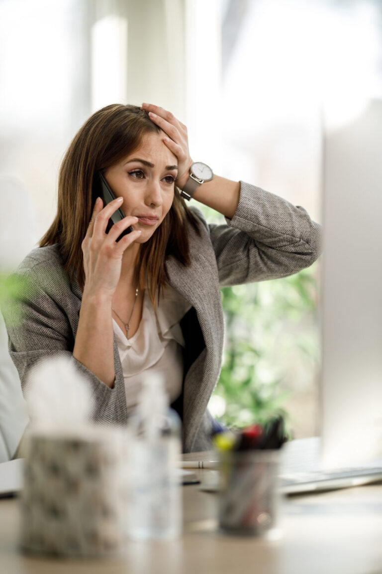 Uma mulher de blazer cinza parece estressada, possivelmente lidando com 14 erros ao contratar uma operadora, segurando a cabeça com uma mão enquanto fala em um smartphone, sentada em uma mesa em um ambiente de escritório.