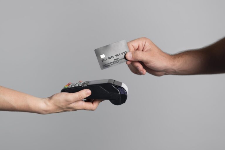 Uma pessoa segura um terminal de pagamento enquanto outra pessoa segura um cartão de crédito acima dele, pronta para fazer um pagamento sem contato - agora possível, pois as lotéricas vão aceitar cartões de crédito - contra um fundo cinza claro.