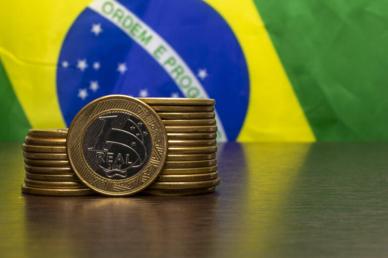 Uma pilha de moedas de real fica em uma mesa com a bandeira do Brasil ao fundo, simbolizando as discussões que os estados e o congresso planejam criar um imposto no Brasil.