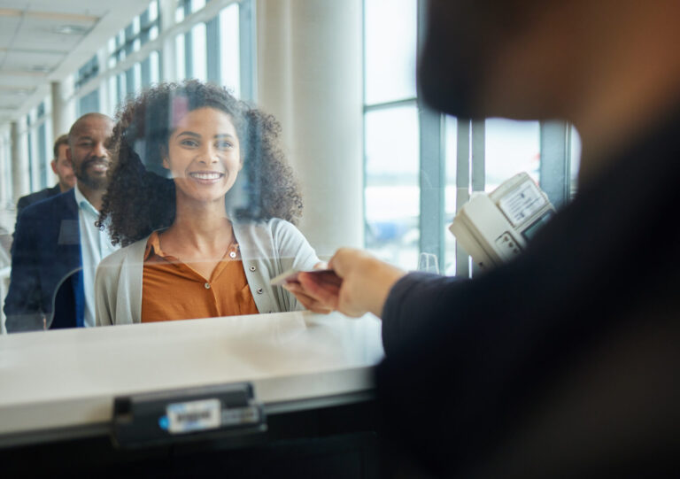 Uma mulher sorri ao entregar seu passaporte a um funcionário da companhia aérea em um balcão de check-in no aeroporto, com um homem na fila atrás dela, mostrando como um lotérico cria material para aproximar o cliente durante a experiência de viagem.